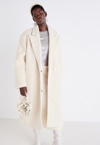 Manteau en laine crème avec grands revers, deux poches avant et fermeture à boutons. Associé à un haut argent clair, une jupe texturée et un sac en satin.