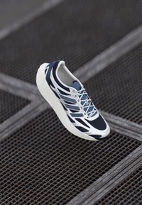 Hvide og marineblå atletiske sneakers med mesh og syntetisk overlay, svævende over en metalgitteroverflade.