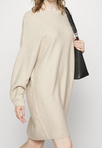 Beige pletené šaty s dlouhými, volnými rukávy. Hladký vzor a lodní výstřih. Modelka drží černou tašku s texturovaným popruhem.