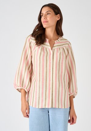 Mujer de cabello largo y castaño que lleva una blusa suelta de rayas verticales en rosa, beige y blanco, abotonada, y pantalones vaqueros azul claro.