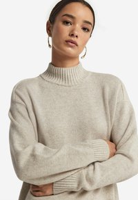 Sweter dzianinowy w jasnym beżu z wysokim, prążkowanym kołnierzem. Posiada obniżone ramiona i prążkowane mankiety, co dodaje faktury.