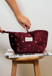 Burgundowa welwetowa kosmetyczka z różowym haftem kwiatowym, zamknięcie na zamek, oraz tkaninowa metka z napisem "The Flat Lay Co." na stołku z magazynami.