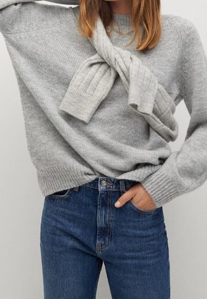 Person iført en grå strikket sweater og jeans, med en matchende grå sweater draperet og bundet omkring skuldrene.