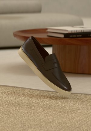 Tamno smeđa kožna mokasina s kremastim đonom lebdi iznad bež teksturiranog tepiha, drvenog okruglog stola i složenih knjiga u pozadini.