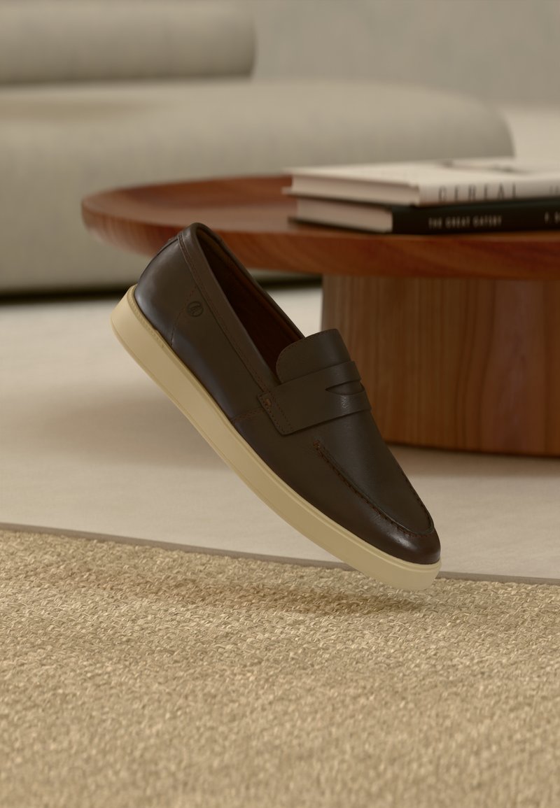 Καφέ σκούρο δερμάτινο παπούτσι τύπου loafer με κρεμ σόλα αιωρείται πάνω από μπεζ υφασμένο χαλί, ξύλινο στρογγυλό τραπέζι και στοιβαγμένα βιβλία στο φόντο.