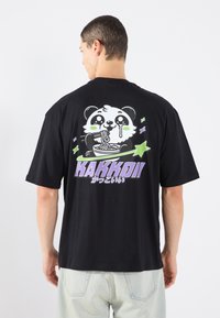 Sort bomulds t-shirt med et tegneseriepanda, der spiser nudler, og teksten "KAKKOII" i lilla. Korte ærmer og afslappet pasform.