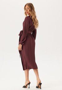 Robe plissée bourgogne à manches longues et à taille ceinturée, atteignant le mollet. Tissu lisse avec des plis verticaux et une coupe décontractée.