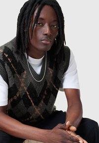 Gilet tricoté en vert foncé avec un motif de diamant noir et orange ; associé à un T-shirt blanc et un collier en chaîne argenté.