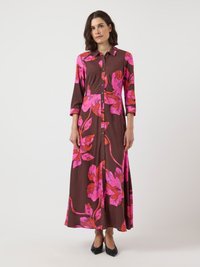 Kvinna i en brun maxiklänning med stora rosa och röda blommönster, knäppning framtill, trekvartsärmar och svarta spetsiga klackar.