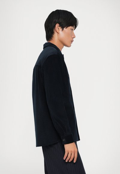 Tumesinine corduroyst särk, millel on nööbiga kinnine ja pikad varrukad, kontrastsete õlavarre paneelidega. Sile tekstuur ja mugav lõige, külgvaade.