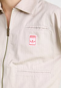Šviesiai smėlinis zip-up džemperis su baltais vertikaliais dryžiais, turintis krūtinės kišenę ir raudoną Adidas logotipo pleistrą. Lygi audinio tekstūra.