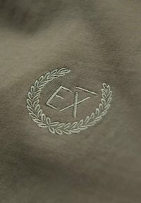 Olivgrüner Stoff mit einem gestickten Emblem von Lorbeerblättern, die "EX" in heller Fadenfarbe umgeben, mit einer glatten Textur.