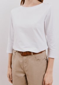 Top bianco a maniche lunghe con bottoni sulle spalle, abbinato a pantaloni beige e una cintura marrone. Tessuto liscio con un taglio classico e una vestibilità comoda.