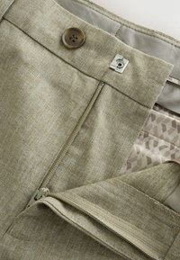 Close-up of beige trousers waistband with button, zipper, and metal hook fastening, showing fabric texture and inner lining pattern.
