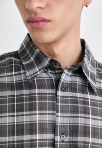Camicia a quadri grigia e nera con colletto classico, apertura con bottoni e texture liscia, caratterizzata da linee bianche contrasta su tutto il tessuto.