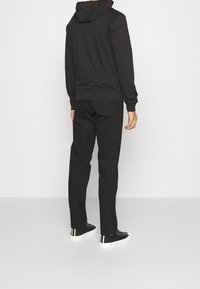 Svart hoodie med en tight passform och huva, tillsammans med svarta byxor och svarta sneakers med vita detaljer och en strukturerad vit sula.