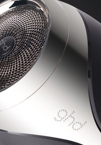 Sèche-cheveux avec un design métallique argenté et noir. Caractérisé par une grille frontale en mesh, une forme circulaire et une surface texturée avec des accents de logo.