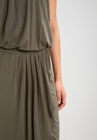 Olive green dress made of soft fabric with a loose bodice and gathered waist, featuring vertical pleats and an elastic waistband.