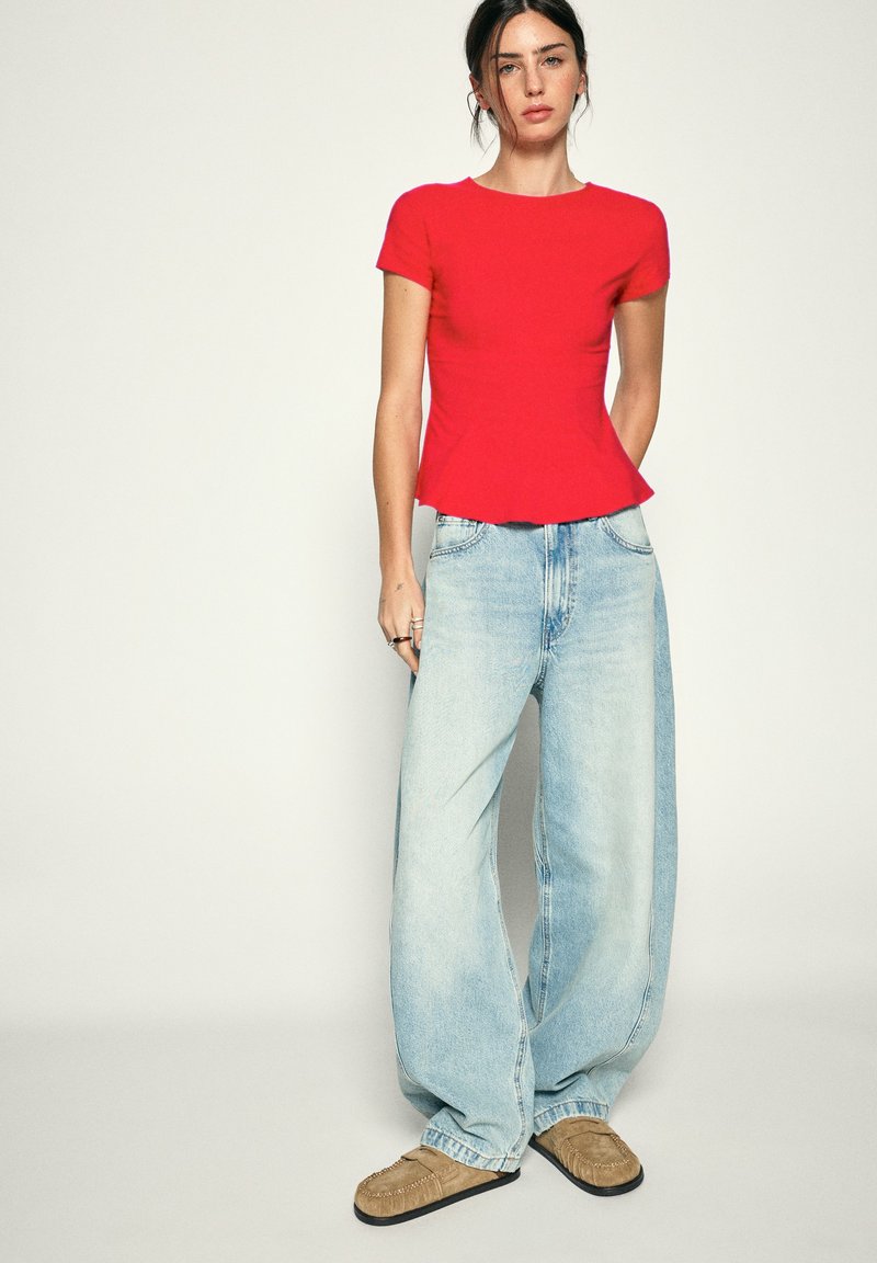 Femme debout portant un haut rouge à manches courtes ajusté, un jean bleu clair à jambes larges et des mocassins couleur sable sur un fond uni.