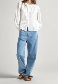 Camicia bianca a maniche lunghe con texture e righe verticali, abbinata a jeans a gamba larga blu chiaro e sandali con plateau marroni.