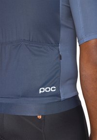 Sötétkék kerékpáros mez rövid ujjakkal, testhezálló fazonban, diszkrét cipzáras zsebbekkel és fehér "poc" logóval az oldalon.