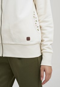 Kremfarget zip-up hettegenser med sidelommer, med grønne og rosa broderte plante-design og et brunt logo-lapp nederst.