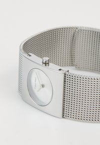 Srebrni Calvin Klein sat s mrežastim remenom i pravokutnim minimalističkim licem koje prikazuje 10:11 na bijeloj pozadini.
