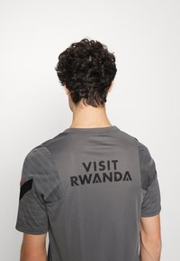 Grå idrottströja med korta ärmar, med texturerade axelpaneler och "BESÖK RWANDA" tryckt med fet svart text på baksidan.