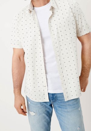 Mann trägt weißes Kurzarmhemd mit kleinen schwarzen Mustern über einem schlichten weißen T-Shirt und hellblauen zerrissenen Jeans.