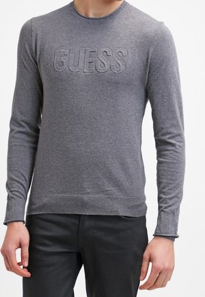 Pull gris à manches longues avec logo "GUESS" embossé, porté avec un pantalon noir par une personne debout, les mains détendues le long du corps.