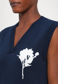 Marineblå ermeløs genser med dyp v-hals, med et hvitt blomstermønster på brystet. Strikket tekstur med ribbede kanter.