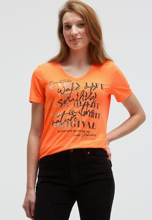 V-neck, short-sleeve t-shirt in bright orange cotton, featuring printed black and white text in varying fonts and sizes. Paired with black jeans.