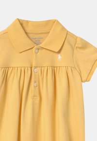 Žluté polo šaty s límečkem, krátkými rukávy a plisovanou sukní. Obsahují tři knoflíky a malé vyšívané logo.