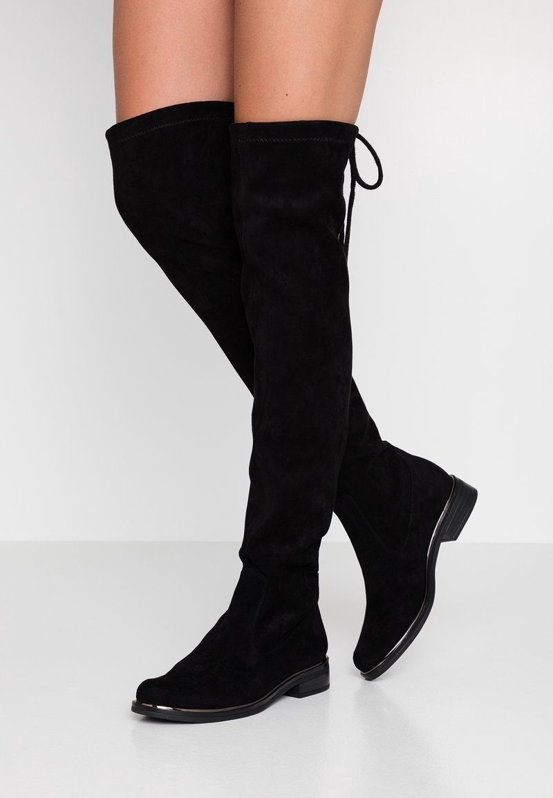 Jambes portant des bottes hautes en daim noir avec des talons bas et un détail de nœud à l'arrière, debout sur un sol blanc contre un mur blanc.
