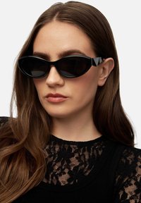 Black cat-eye sunglasses with dark lenses, wide geometric frames, and triangular accents on the temples. Worn over a black lace top.