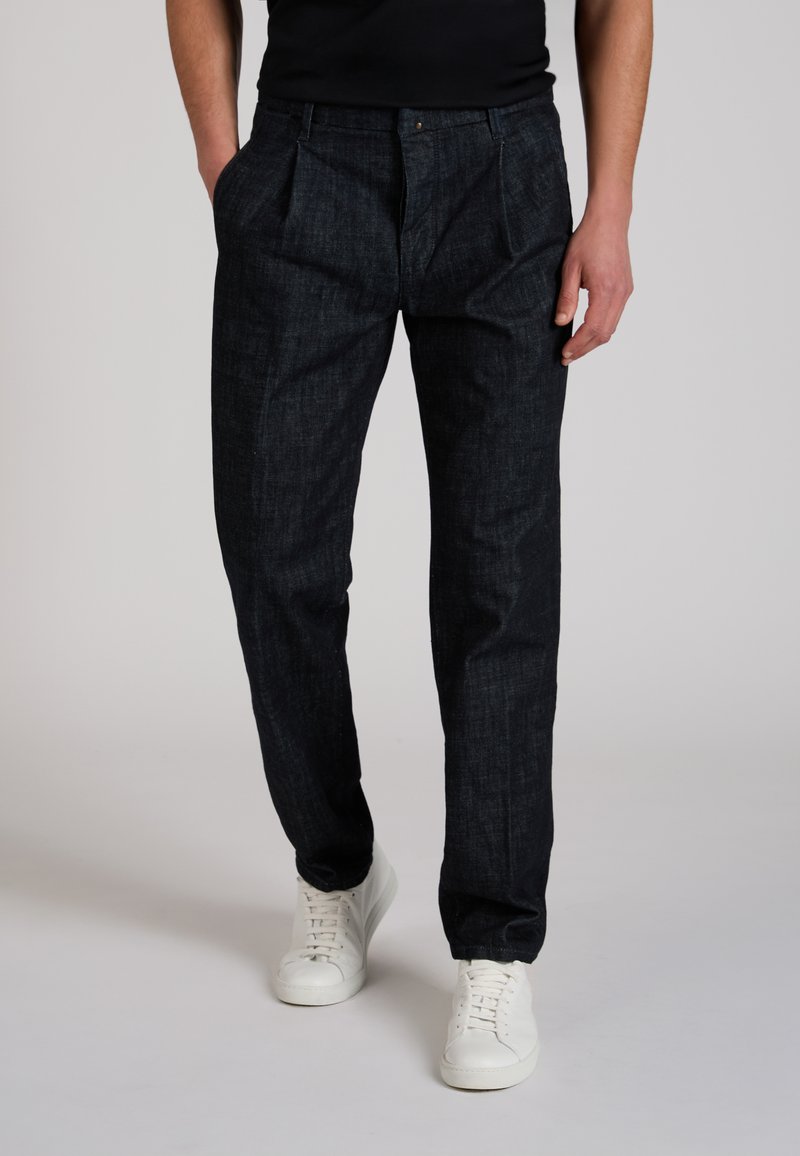 Man standing wearing dark blue pleated trousers, black top, and white sneakers on a plain light background.