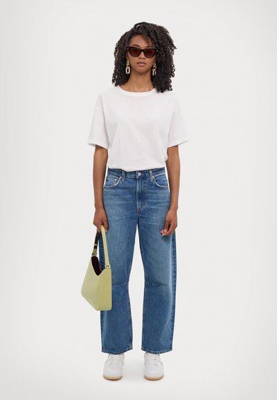 Weißes oversized T-Shirt, blaue weit geschnittene Jeans, weiße Sneaker und eine hellgrüne Schultertasche. Zu den Accessoires gehören dunkle Sonnenbrillen und Ohrringe.