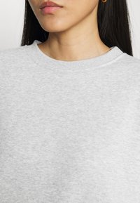 Primer plano de la parte inferior de la cara y el cuello de una mujer que lleva un suéter de cuello redondo gris claro y un pequeño aro de oro.