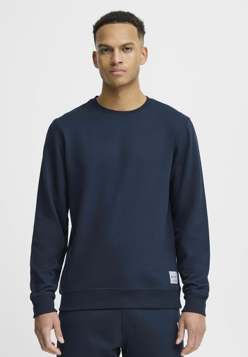 Mornarsko modra majica s crewneck iz mešanice mehke bombažne tkanine z rebrastimi manšetami in robom, s poudarjeno majhno etiketo na spodnji strani.