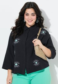 Zwarte textuur blouse met geborduurde oogmotieven, gecombineerd met mintgroene broek en een sprankelende gouden handtas.