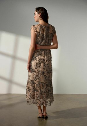 Nő beltéren áll, bézs ruhát visel fekete virágos csipke rávarrással, fodros ujjakkal és bokapántos magassarkúval, háttal néz.