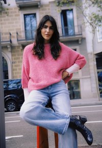 Pull rose flou, coupe décontractée, avec poignets retroussés, associé à un jean bleu clair et des bottines noires. Décor urbain avec des bâtiments en arrière-plan.