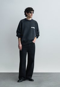 Uomo con capelli ricci che indossa occhiali da sole neri, felpa nera con testo bianco, pantaloni neri a gamba larga e scarpe nere, in piedi contro un muro semplice.