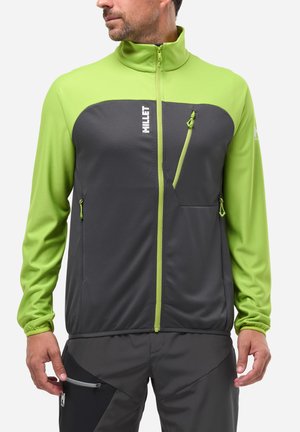Homme portant une veste Millet zippée verte et grise avec des poches poitrine et latérales zippées, associée à un pantalon gris foncé.
