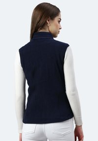 Marineblauer Fleece-Weste mit hohem Kragen, ärmellosem Design und glatter Textur, getragen über einem weißen, gerippten Langarmoberteil, kombiniert mit weißen Jeans.