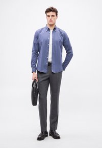 Svetlomodrá košeľa s golierom na gombíky, biele spodné tričko, sivé nohavice, čierny opasok, drží čiernu tašku a čierne topánky. Jednoduchý, na mieru ušitý dizajn.