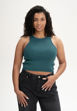 Vrouw met krullend haar, gekleed in een teal mouwloze cropped top en zwarte spijkerbroek, staand met één hand in haar zij en glimlachend.