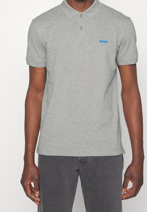 Mann trägt ein hellgraues Poloshirt mit kurzen Ärmeln und blauem "BOSS"-Logo auf der Brust sowie dunkelgraue Hose, steht vor einfarbigem Hintergrund.