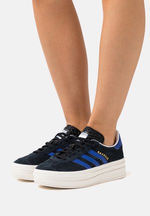 Jambes portant des baskets Adidas Gazelle noires avec des rayures bleues et des semelles épaisses blanches, debout sur un fond blanc uni.