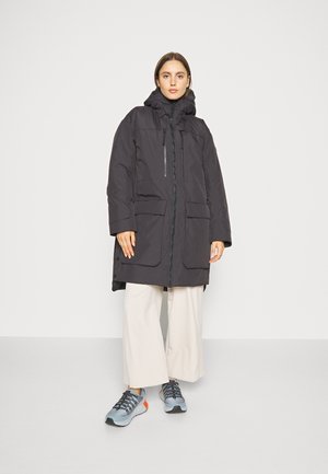 Zwarte gewatteerde jas met een hoge kraag, twee voorkoeken en een rits aan de voorkant. Draagt over lichtgekleurde wijde broek en grijze sneakers.
